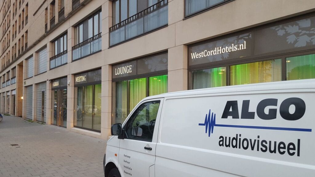 White van with the ALGO audiovisueel logo parked on a city sidewalk in front of storefronts labeled WestCordHotels.nl, Lounge, and Meeting.