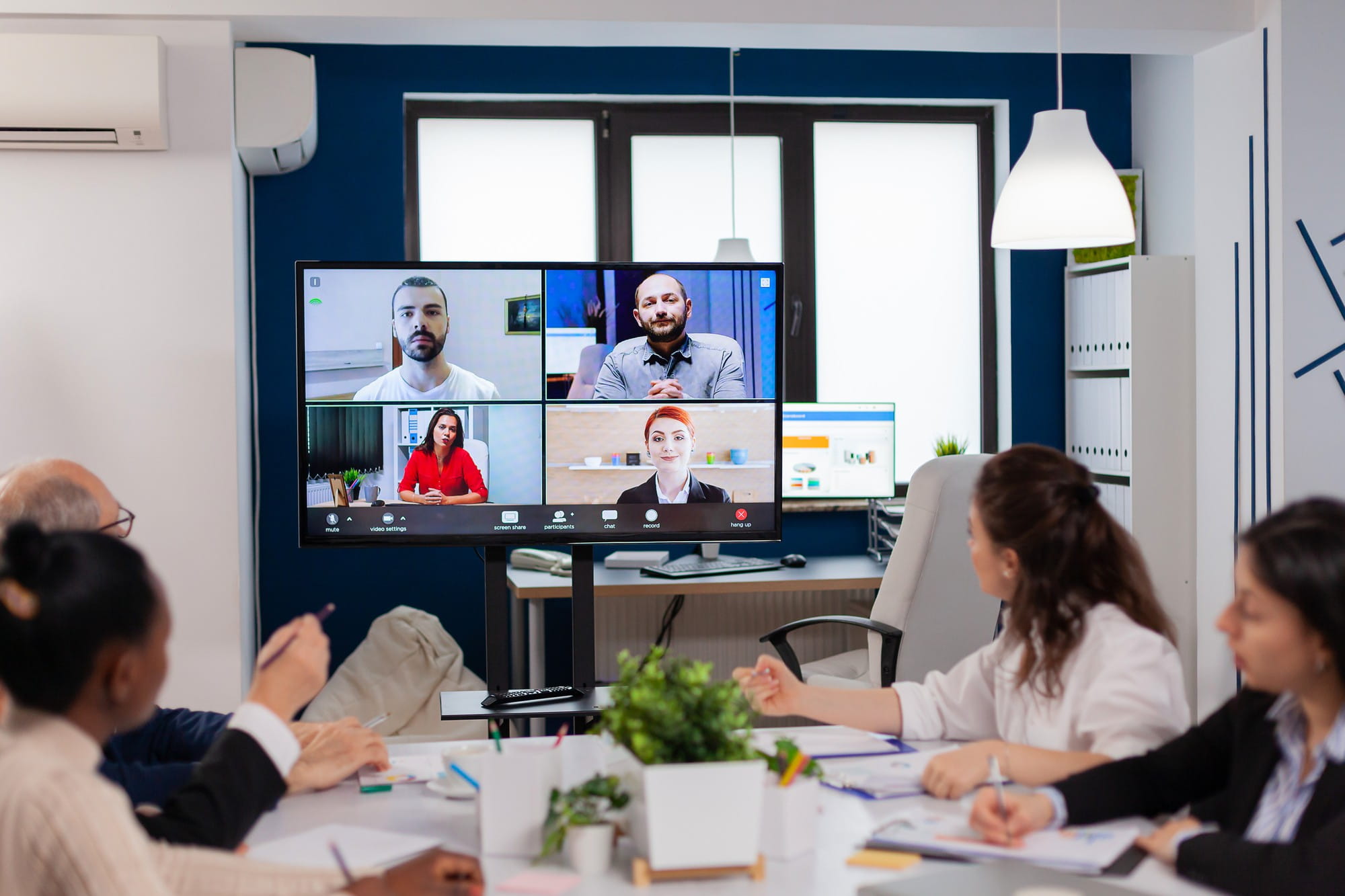 Business meeting in a modern conference room; a large screen shows a six-person video conference.